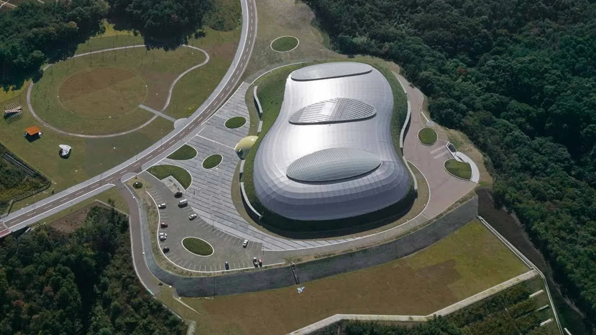 Aerial view of the Bourbon Beans Dome in Miki City, Japan, a curved metal roof set into a green hillside with a park and running track around it